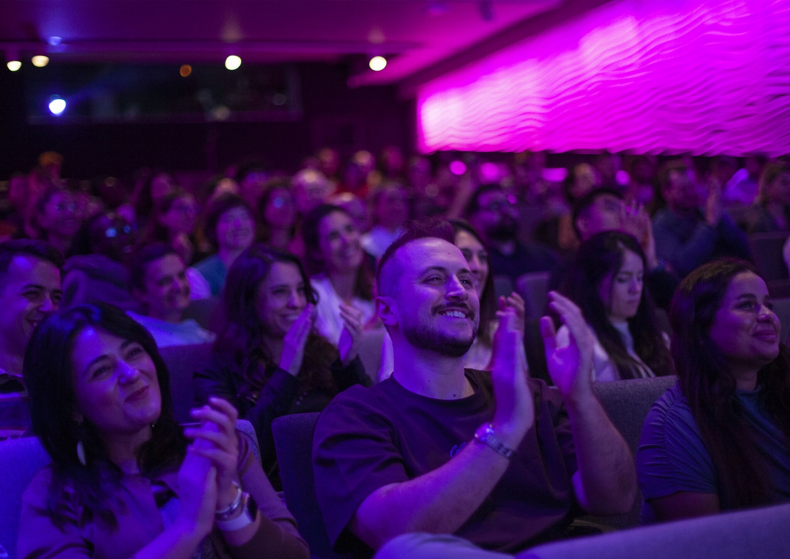 Sami smiling and applauding in a crowded auditorium lit in pink and violet.